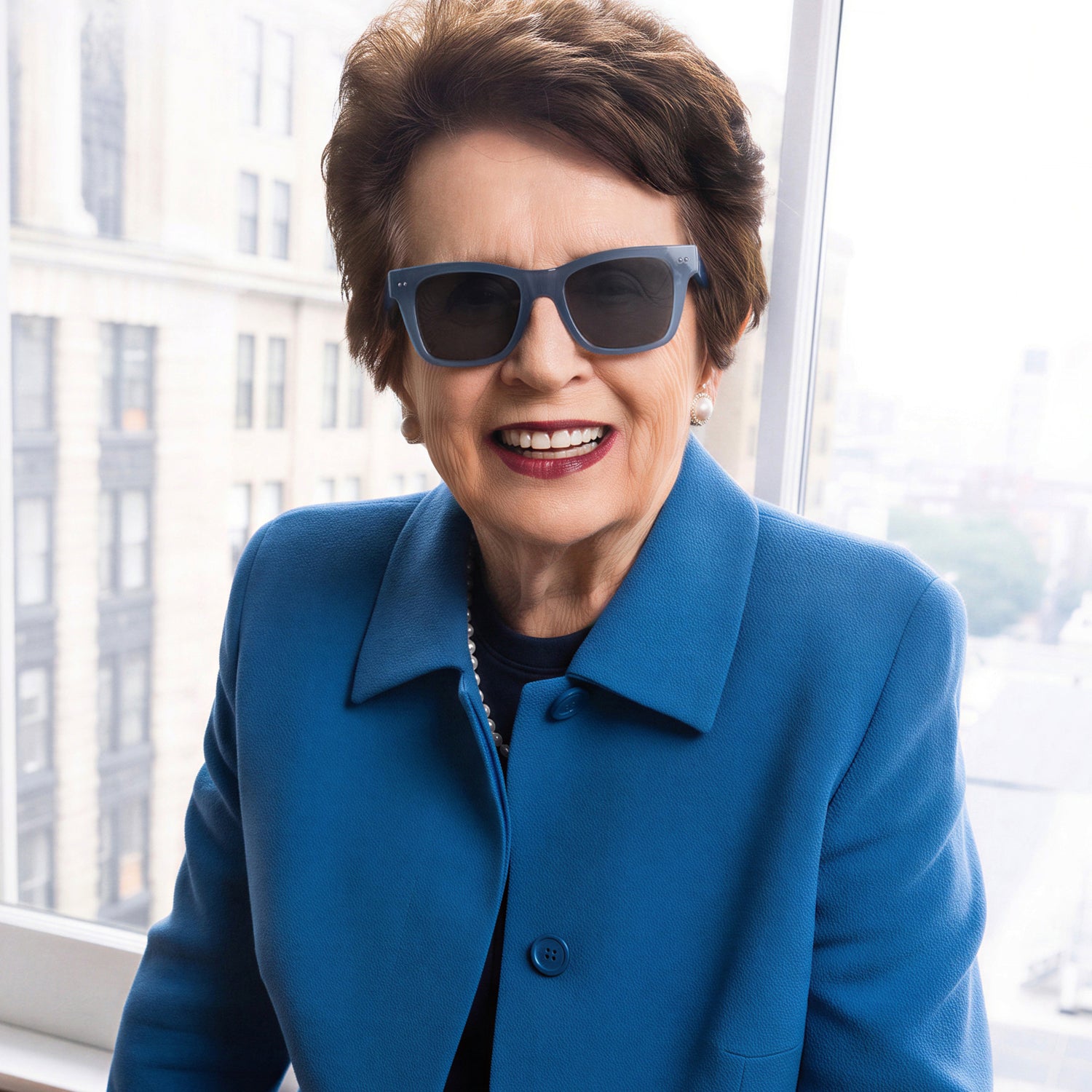 Smiling woman wearing dark blue sunglasses and a blue coat indoors.