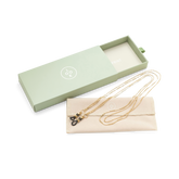 A gold eyewear chain with black end loops rests on a beige pouch beside an open pale green box.