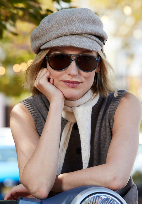 Woman wearing dark sunglasses, a gray cap, and scarf outdoors.