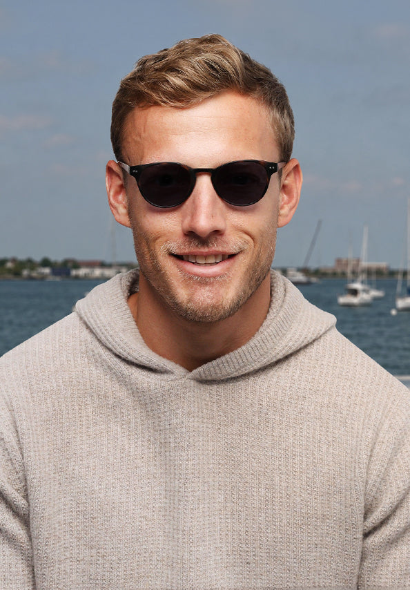 Man wearing black sunglasses and a beige hooded sweater by the water.