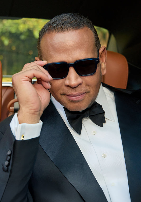 Man in a black tuxedo adjusting dark rectangular sunglasses inside a car.