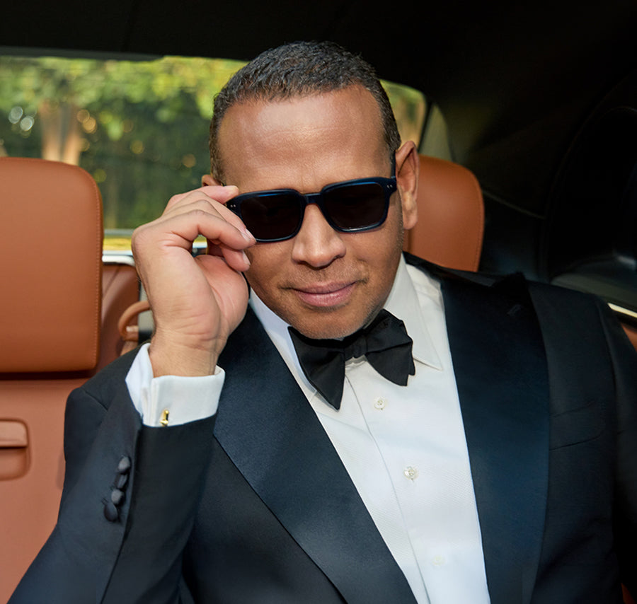 Man in a black tuxedo adjusting dark rectangular sunglasses inside a car.
