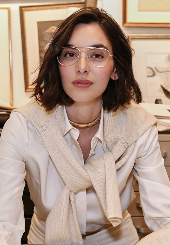 Woman wearing clear gold-rimmed glasses, a white shirt, and a beige sweater draped over her shoulders.