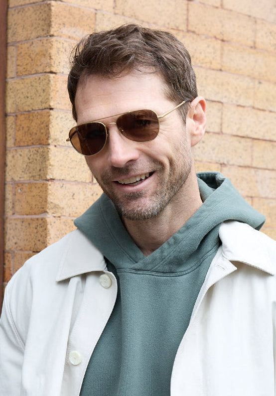 Man wearing brown aviator sunglasses, a green hoodie, and white jacket outdoors.