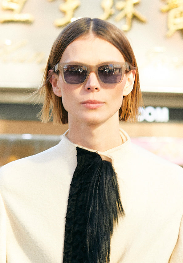 Woman wearing dark rectangular sunglasses and a cream top with a black tie detail.