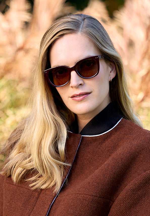 Woman with long blonde hair wearing dark brown sunglasses and a brown coat outdoors.