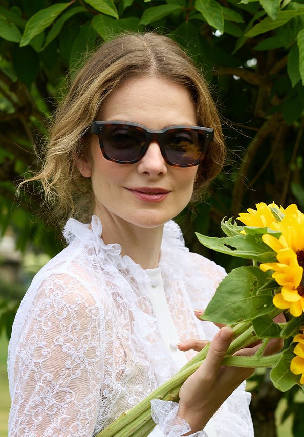 Woman wearing dark sunglasses and a white lace blouse holds a bouquet of bright yellow flowers.