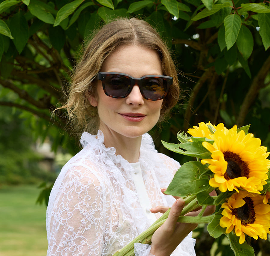 Woman wearing dark sunglasses holds a bouquet of bright sunflowers outdoors.
