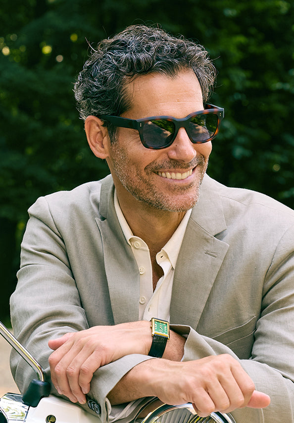 Smiling man wearing dark sunglasses and a beige blazer outdoors.