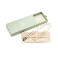 
A gold eyewear chain with black end loops rests on a beige pouch beside an open pale green box.