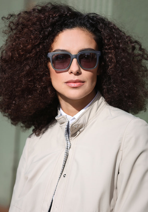 Woman with curly hair wearing oversized gray sunglasses and a light jacket.