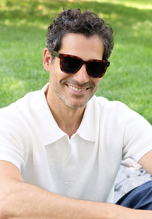 Smiling man wearing dark tortoiseshell sunglasses and a white shirt outdoors.