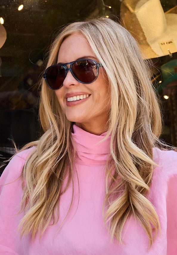 Smiling woman with long blonde hair wearing dark aviator sunglasses and a pink turtleneck.