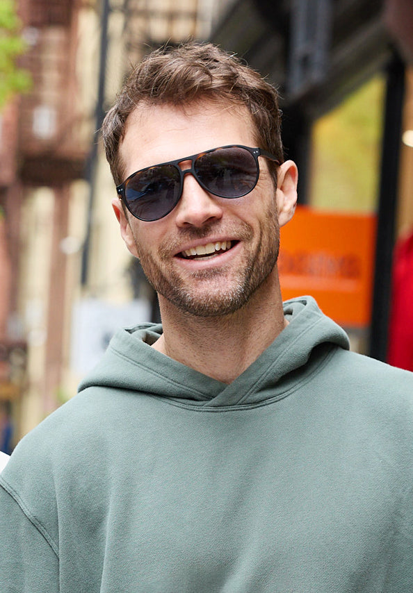 Smiling man wearing black sunglasses and a sage green hoodie outdoors.