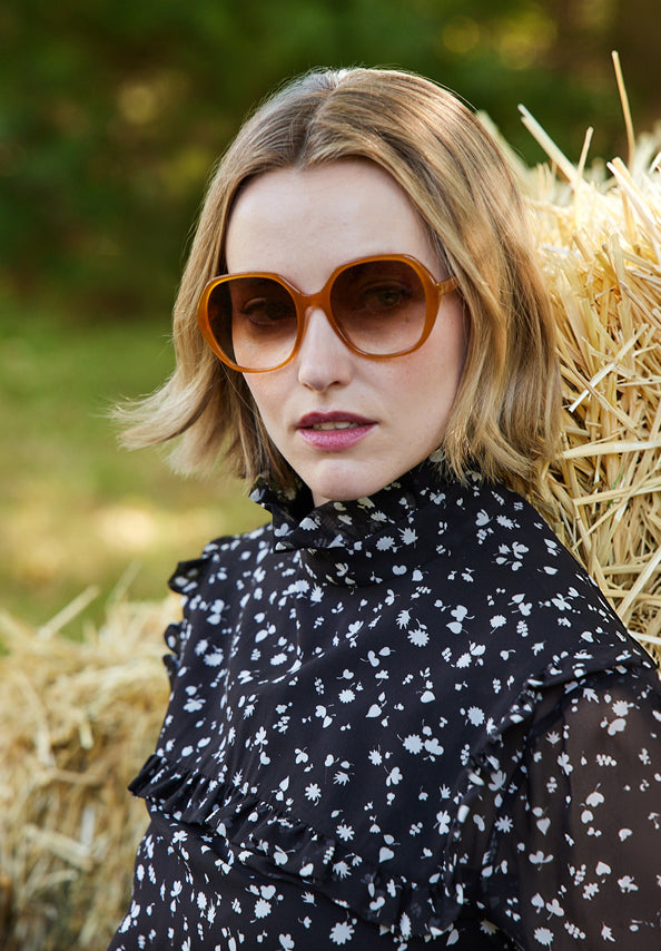Woman wearing oversized amber sunglasses and a black floral blouse beside hay bales.