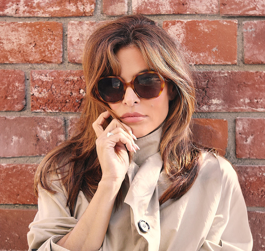 Woman wearing dark sunglasses and a beige jacket posed against a brick wall.