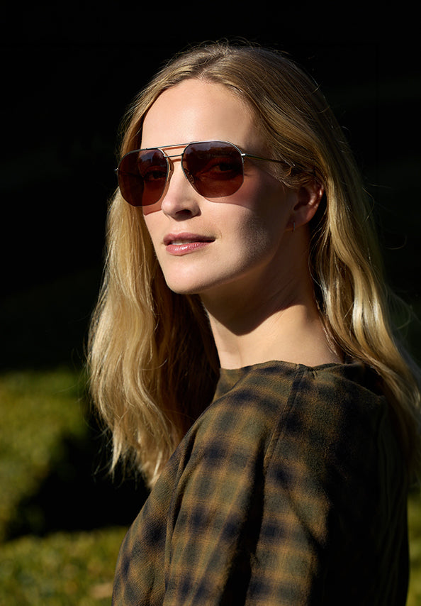 Woman with long blonde hair wearing brown aviator sunglasses and a dark plaid top outdoors.