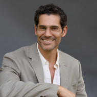 
A person in a dark suit wears silver aviator-style eyeglasses with a double bridge and clear lenses.