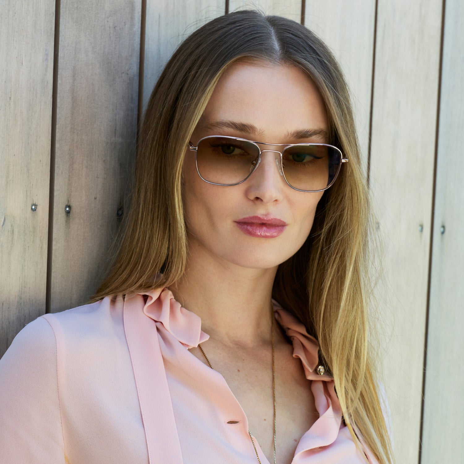 
A person in a pale pink blouse wears shiny rose gold aviator-style eyeglasses with a double bridge and brown gradient tinted lenses.