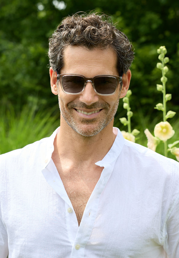 Smiling man wearing brown sunglasses and a white shirt outdoors in a lush garden.