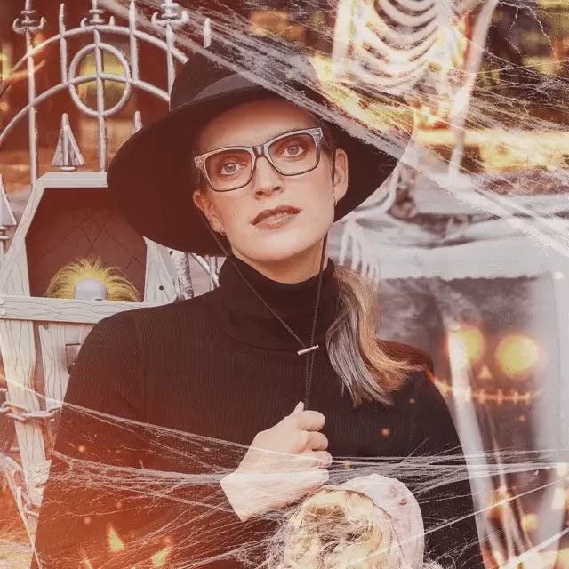 Woman in a black witch hat and LOOK OPTIC glasses among Halloween cobwebs and glowing pumpkins